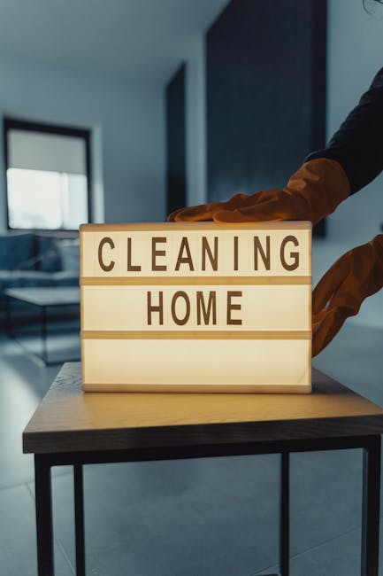 A person wearing an orange glove is placing a wooden block letter sign on a modern, dark-colored table. The sign displays the words 'CLEANING HOME' in bold, black uppercase letters on illuminated pane