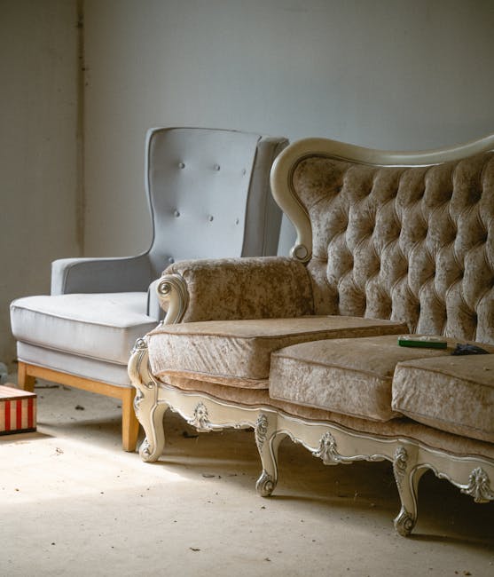 A close-up of a person's hand holding a steam cleaning wand, applying deep cleaning to a grey fabric upholstered armchair. The wand is connected to a professional steam cleaning device, and the nozzle
