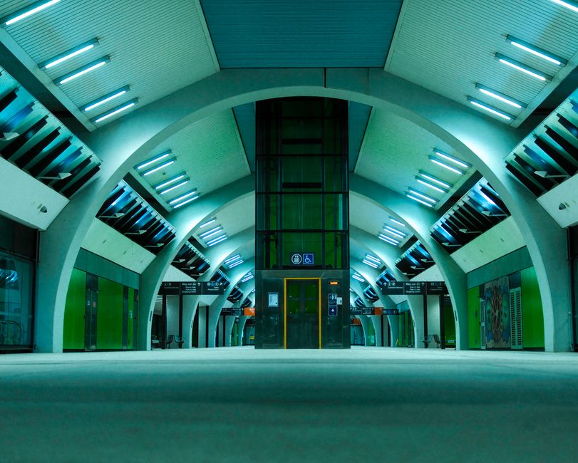 A person with a backpack is descending an escalator inside Isleworth station. The station features contemporary architectural elements with metal supports, glass panels, and tiled flooring. The escala