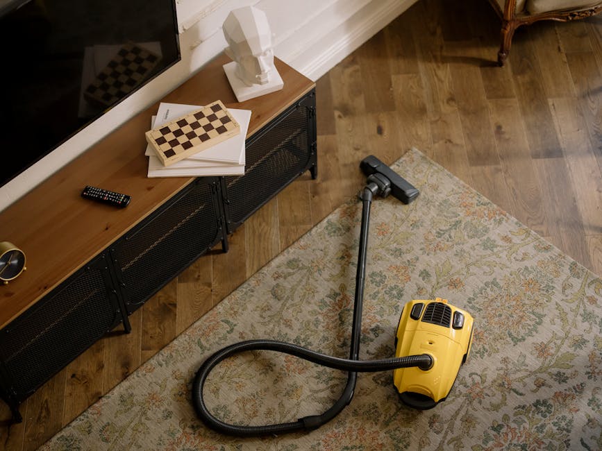 A professional cleaner from Isle Worth Carpet Cleaners is performing deep cleaning on a dark carpeted floor in an indoor auditorium. The cleaner, wearing a light gray jumpsuit and white sneakers, is b